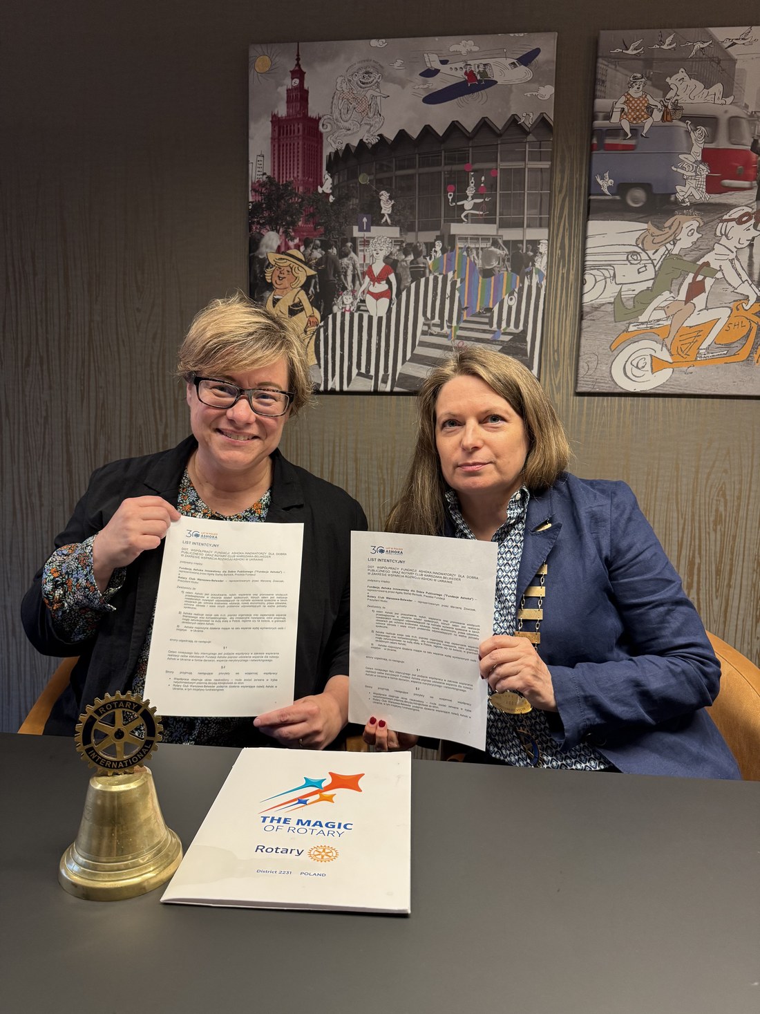 Dwie kobiety przy stole trzymają podpisane dokumenty; obok dzwonek Rotary i tabliczka The Magic of Rotary
