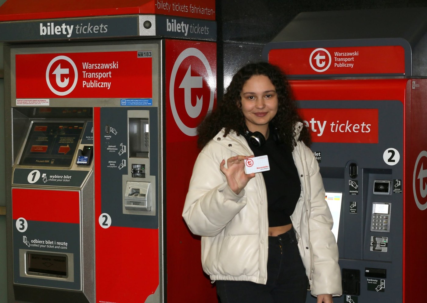 Dziewczyna przy automatach Warszawskiego Transportu Publicznego pokazuje kartę miejską