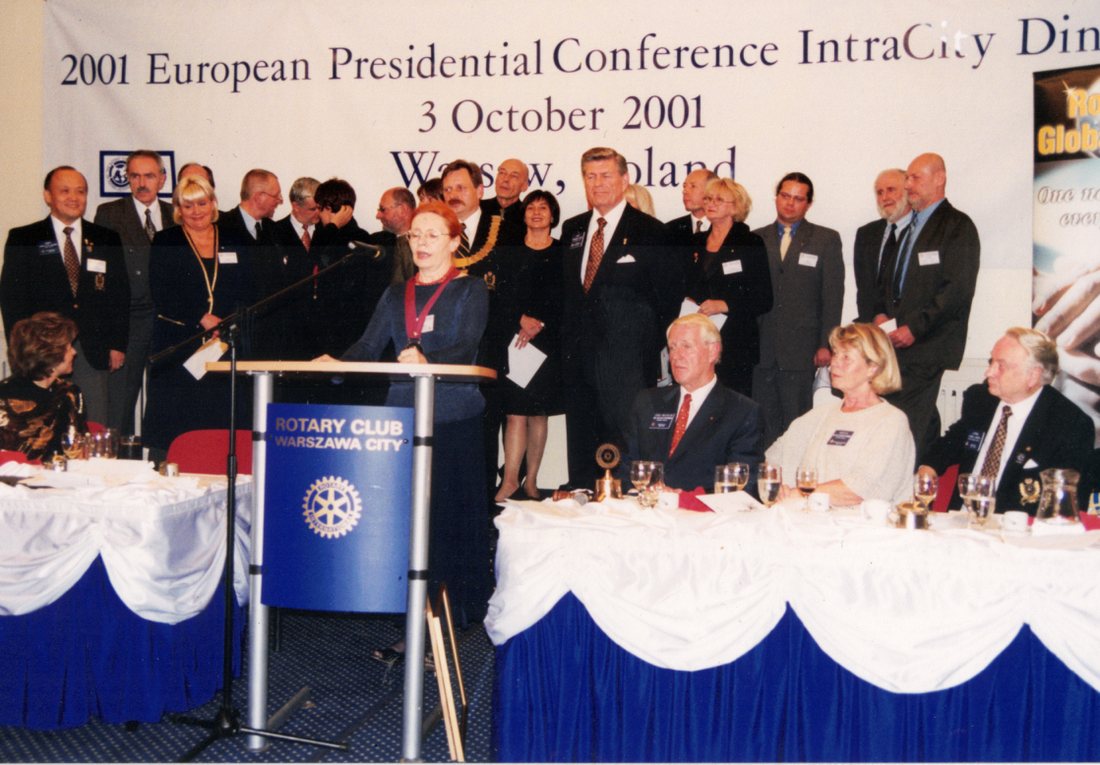Uroczysta kolacja konferencji: kobieta przemawia z mównicy z napisem Rotary Club Warszawa City, za nią rząd uczestników przed banerem „2001 European Presidential Conference IntraCity Dinner”