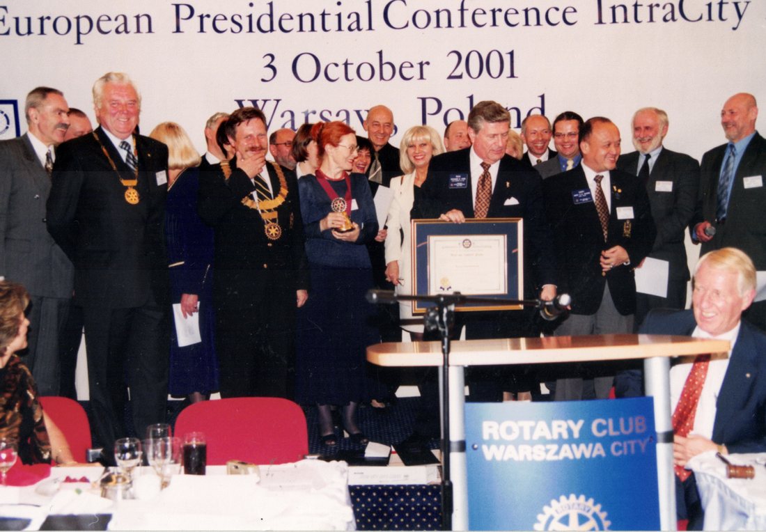Ceremonia wręczenia Karty Klubowej: grupa rotarian przed banerem „European Presidential Conference IntraCity, 3 October 2001, Warsaw, Poland”, pośrodku oprawiony w ramę dokument Karty, przed nimi mównica z napisem Rotary Club Warszawa City