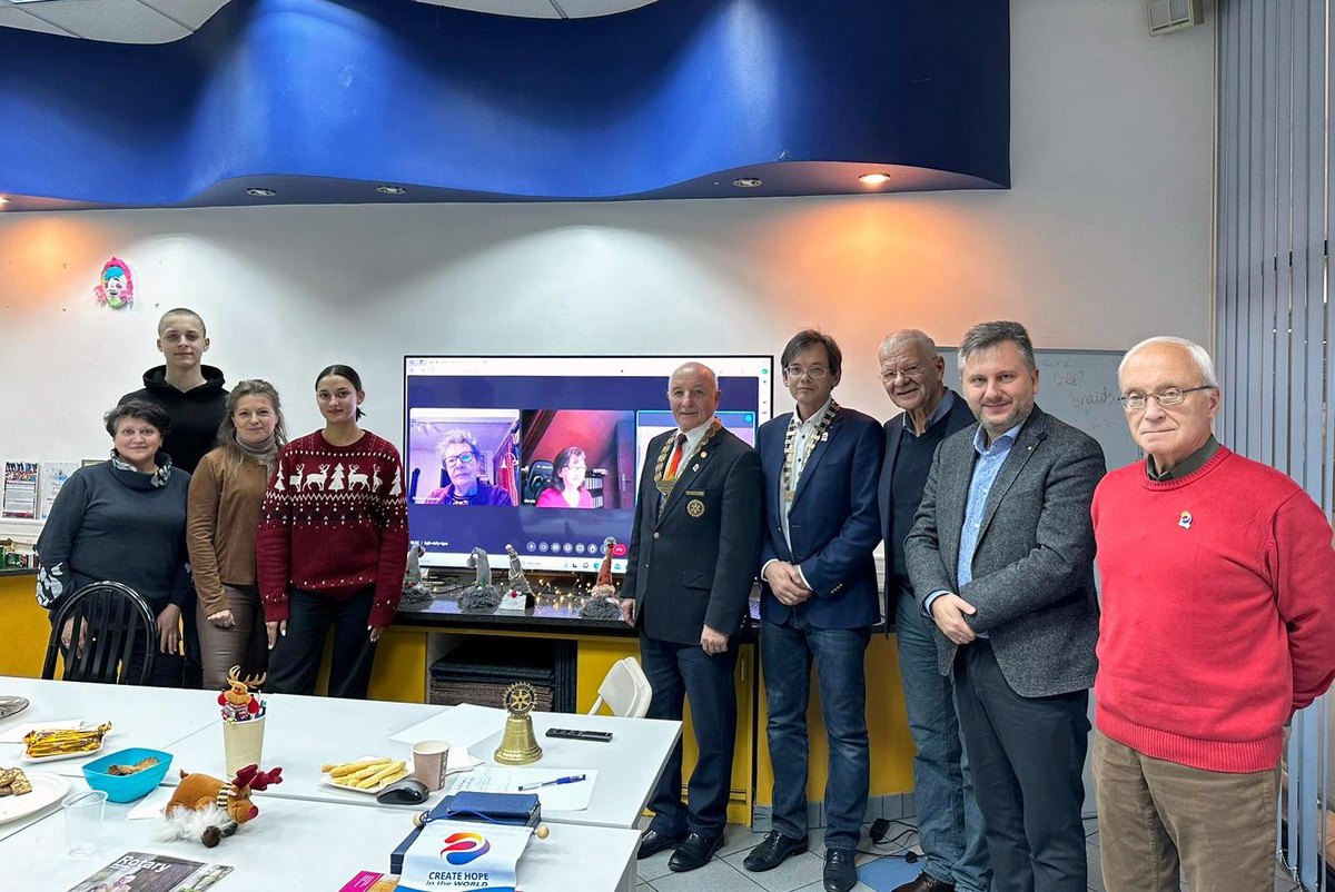 Członkowie i goście klubu przy świątecznym stole; dwaj rotarianie w łańcuchach, na ekranie za nimi uczestniczki spotkania online