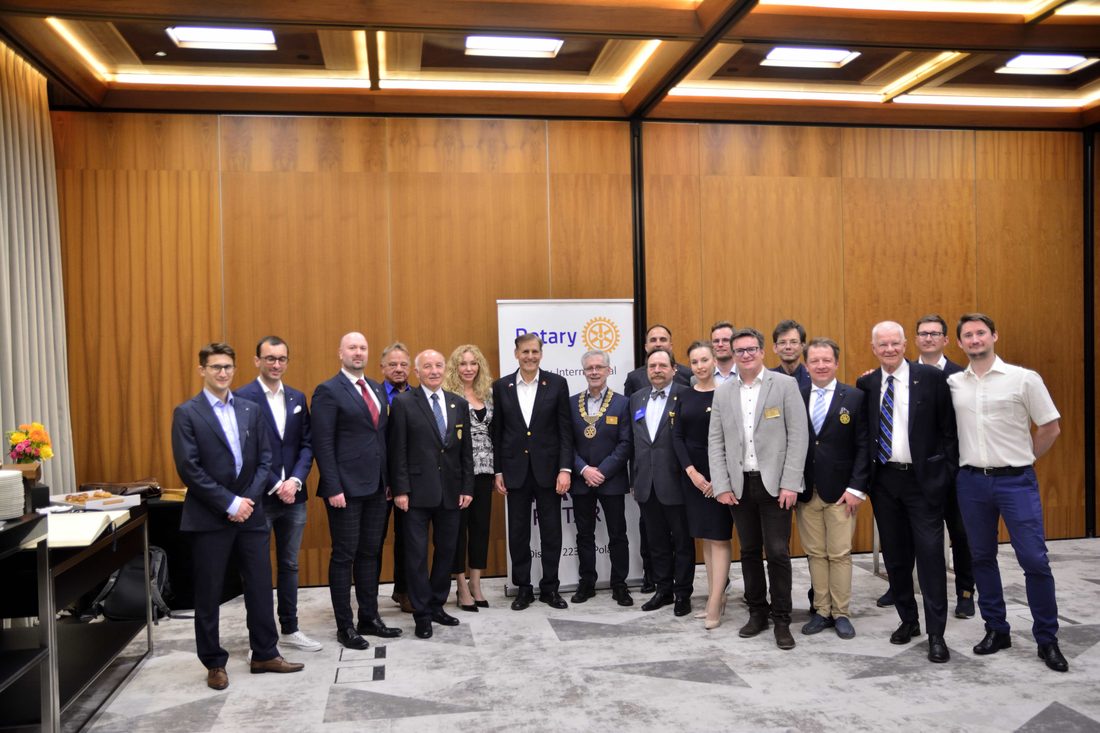 Grupa kilkunastu rotarian pozuje do wspólnego zdjęcia przed roll-upem Rotary International w sali o drewnianych ścianach