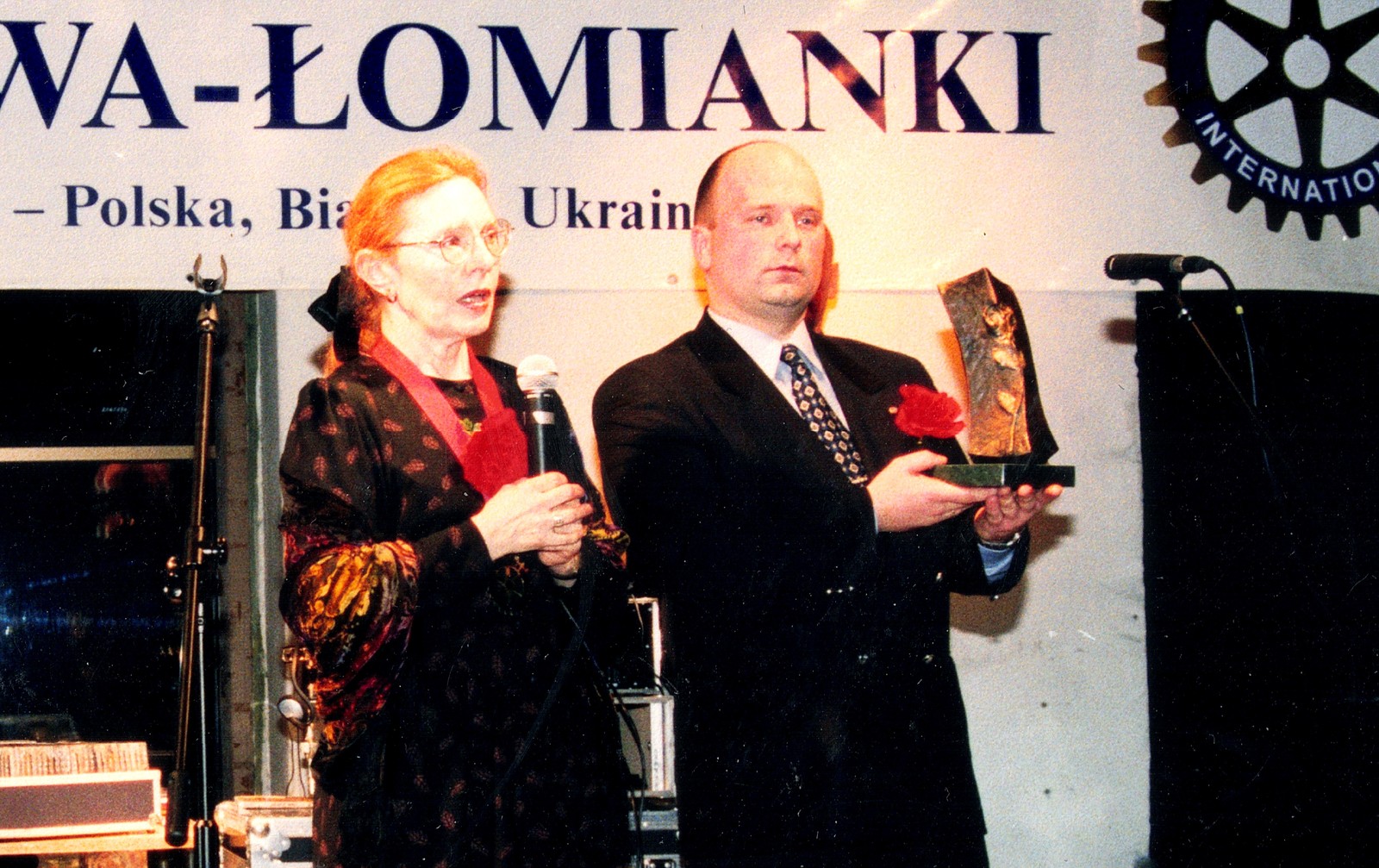 Scena pod banerem „Klub Rotary Warszawa-Łomianki, Dystrykt 2230 – Polska, Białoruś, Ukraina”: kobieta mówi do mikrofonu, obok mężczyzna trzyma statuetkę z czerwoną różą